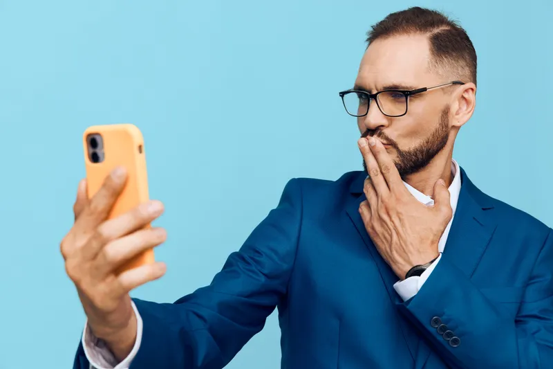 A professional man in a blue suit on an isolated colored background, gazing at a smartphone with a thoughtful expression, conveying focus, strategy, and business mindset.