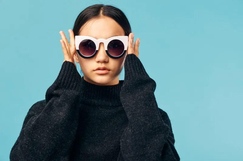 Young woman wearing large white sunglasses and black sweater posing against pastel blue background with serious expression and minimalist style fashion portrait.