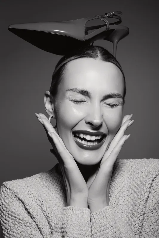 Smiling white woman with light skin and closed eyes wearing a knitted sweater balances a high heel shoe on her head. Concept of joy, fashion, and playful mood in black and white.