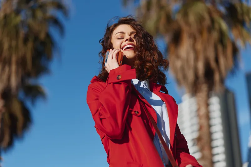 Young woman with curly hair wearing a red jacket talks on a phone outdoors, smiling brightly under a clear blue sky with palm trees and a city backdrop.