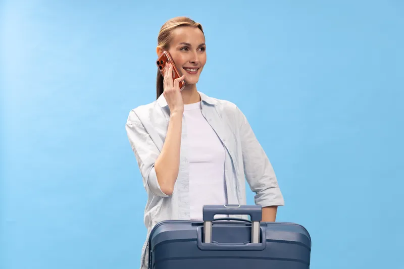 Young woman with suitcase talking on smartphone, smiling and looking aside. Travel concept with luggage and casual outfit on blue background, vacation and trip preparation theme.