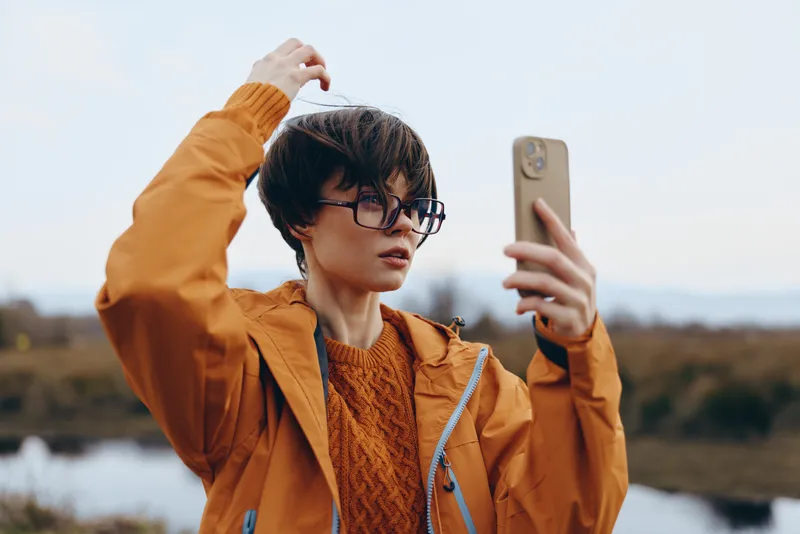 Woman outdoors lifestyle, taking a selfie with phone by the lake, wearing glasses and orange jacket, adjusting hair, calm expression, soft wind, natural light