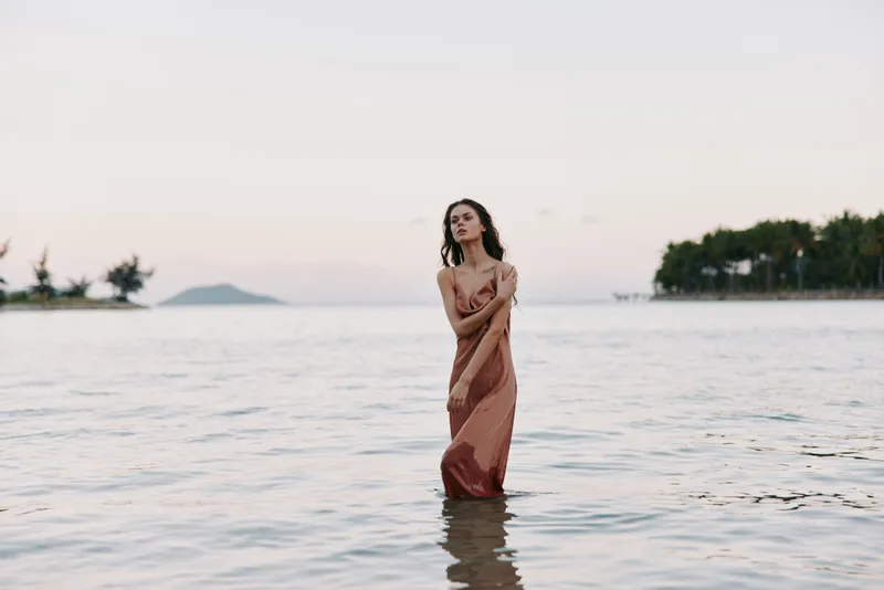 A solitary woman stands in shallow sea water at sunset, wearing a flowing dress, creating a tranquil mood, soft light, reflective surface, and a calm horizon.