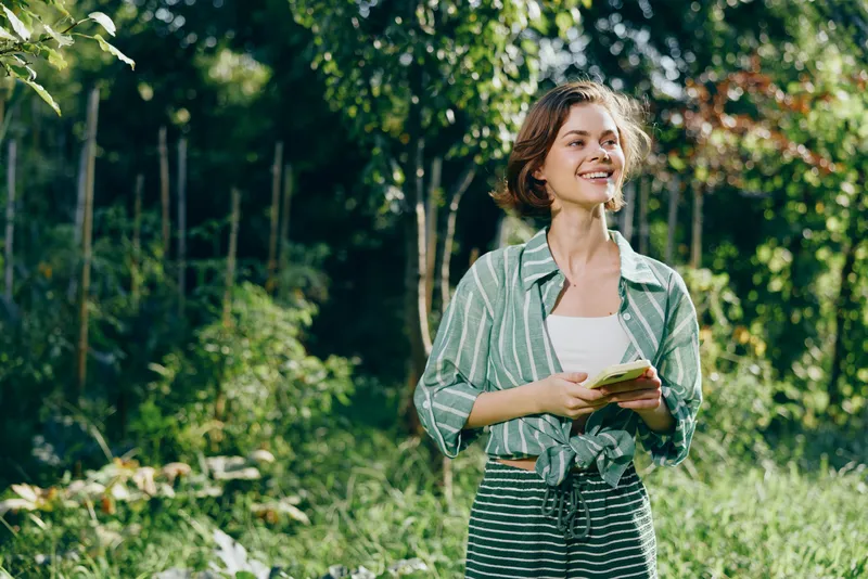 Young woman stands in a sunlit garden, smiling and holding a notebook, capturing a fresh outdoor moment.