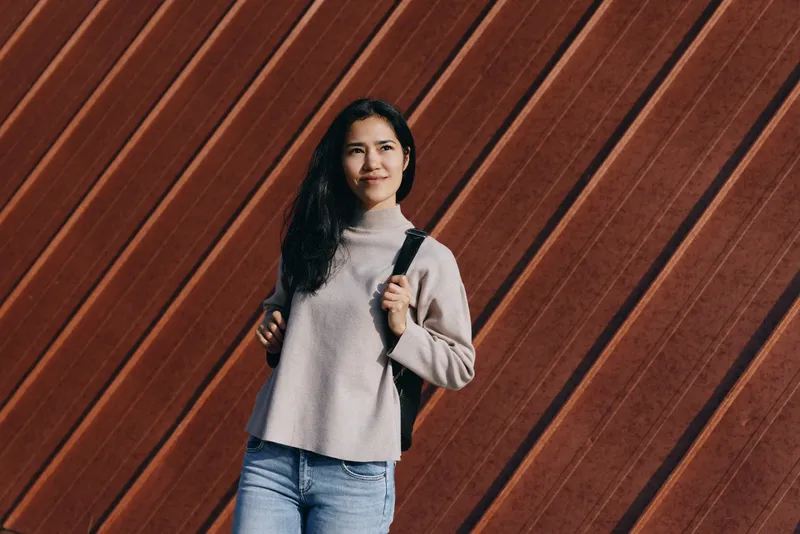 Woman with backpack wearing a cozy sweater and jeans stands before a diagonal wooden wall in an outdoor setting, conveying casual style and confident exploration