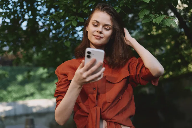 A woman in a red blouse outdoors, looking at a smartphone with a calm expression. A lifestyle scene showing casual, everyday connectivity