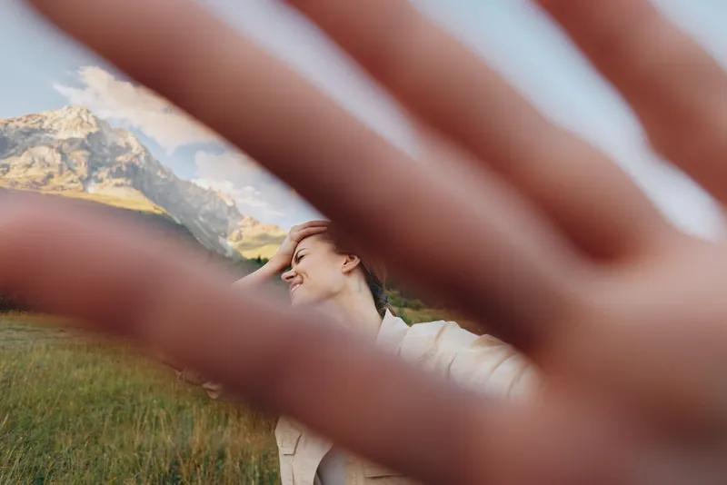 A young woman with brunette hair in a white shirt stands in a grassy field with rugged mountains in the background, smiling softly as a hand frame adds an artistic foreground touch