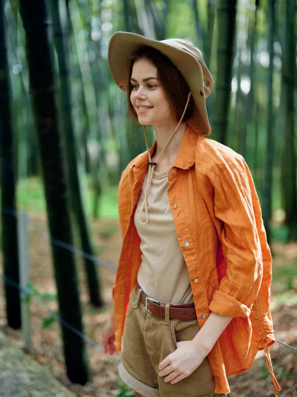 Woman nature bamboo hat hiking smile forest travel portrait of a young female hiker in orange shirt and shorts walking a trail through a serene bamboo grove, enjoying outdoor adventure.