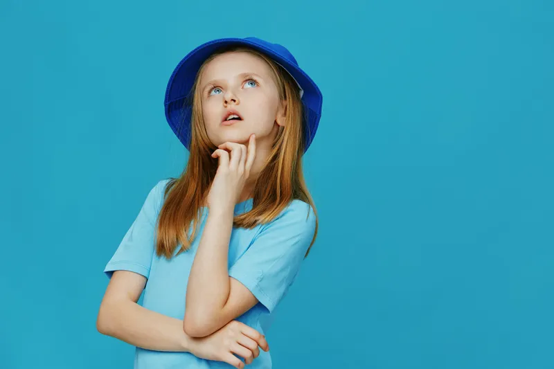 A thoughtful girl in a blue hat and t shirt poses against a vibrant turquoise background, showcasing curiosity and playfulness in a fresh summer vibe