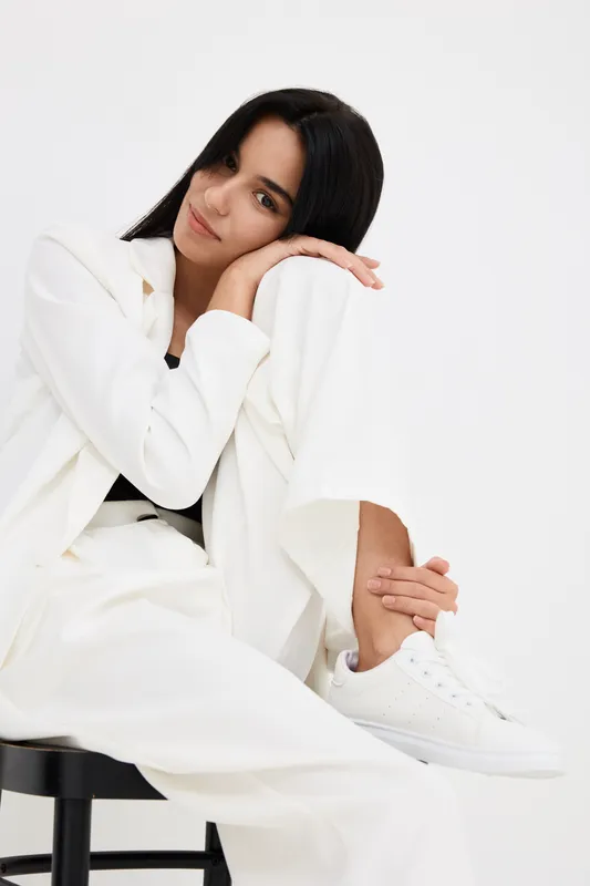 young woman in white suit sitting on black stool, looking relaxed and thoughtful against plain white background, casual outfit with white sneakers