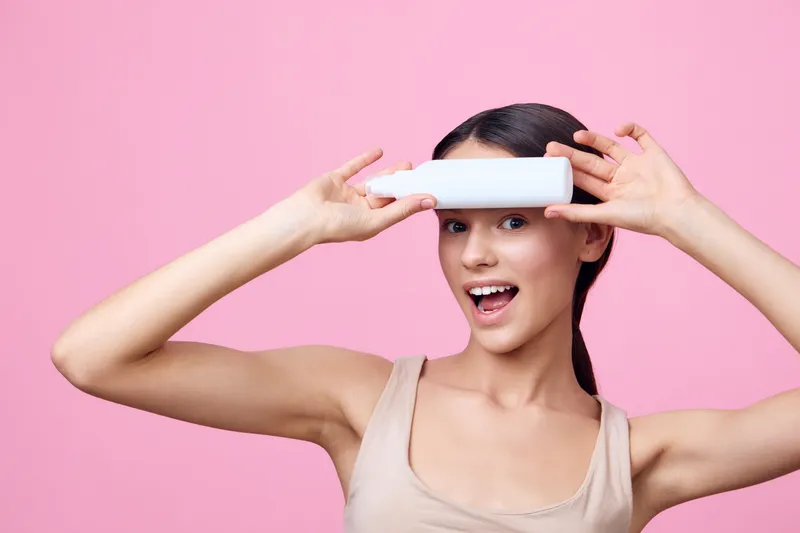 Young smiling woman holding a skincare tube against a pink background, promoting beauty and self care with vibrant colors and a playful mood