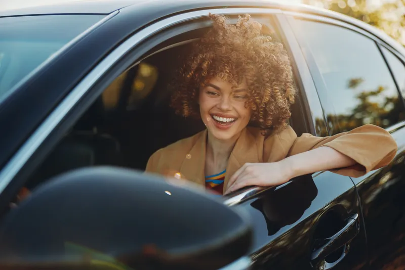 A young woman with curly hair leans out of a car window, smiling softly as warm retro light washes over the scene, conveying a carefree candid moment.