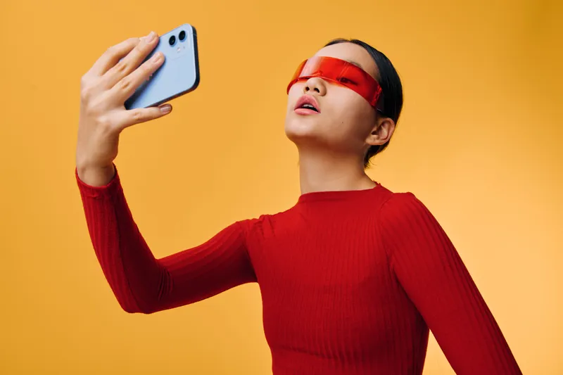 Young woman wearing red translucent sunglasses and red sweater takes a selfie with smartphone against yellow background. Modern fashion, beauty and technology concept with confident style.