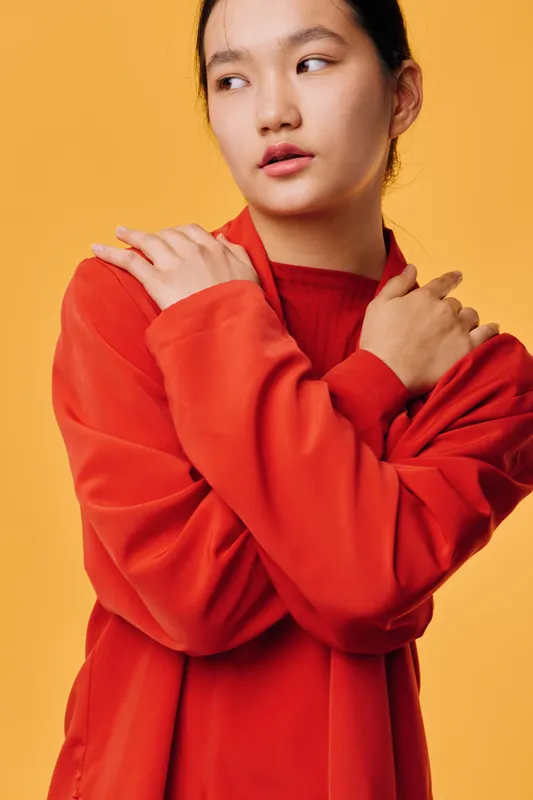 Young woman wearing red sweater posing with crossed arms looking sideways on yellow background. Asian female model with natural makeup in casual style for fashion or lifestyle.