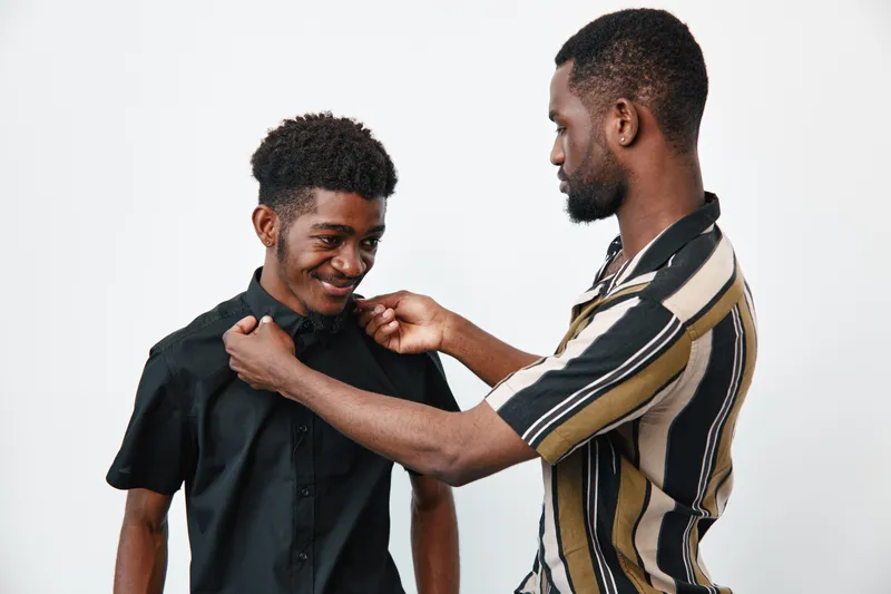 Two african men with dark skin adjusting shirt collar and smiling in studio. Friends, casual clothing and grooming with positive interaction and care between young males.