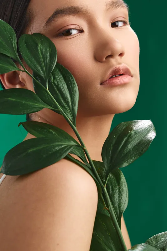 Asian woman closeup portrait with lush plant leaves beside her face in a studio. The image conveys natural beauty, calm mood, and botanical elegance with vibrant green tones.