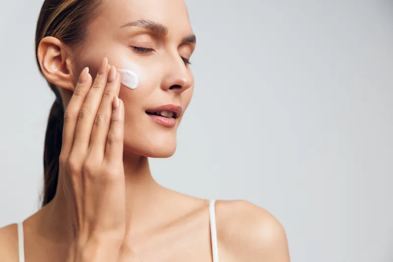 A young woman with closed eyes applies facial cream on her cheek in a bright studio. Concept for skincare, beauty routine, moisturizing, and healthy skin care.