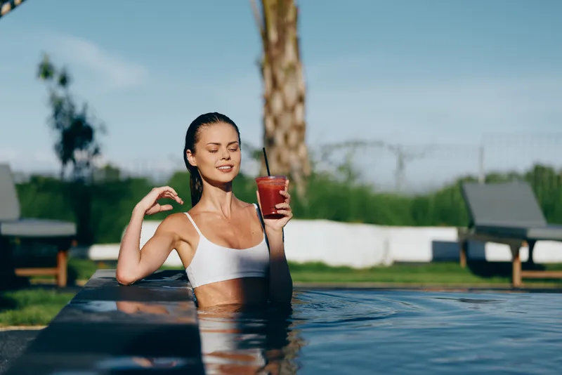 Cheerful woman in a white bikini relaxing in a pool, sunlit moment, holding a glass of juice, relaxed posture, soft breeze, lush greenery backdrop, carefree summertime vibe