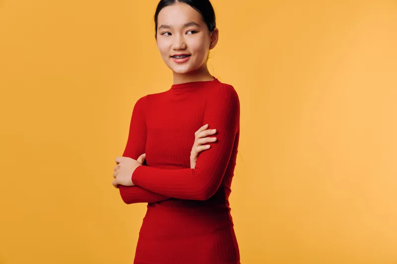 Young woman smiling with crossed arms in red long sleeve shirt against yellow background. Portrait of confident female with casual outfit and natural makeup for lifestyle concept.