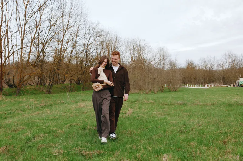 Young couple walks together on a green field with a baby goat in arms under a cloudy sky, enjoying a peaceful moment in casual clothing during early spring.
