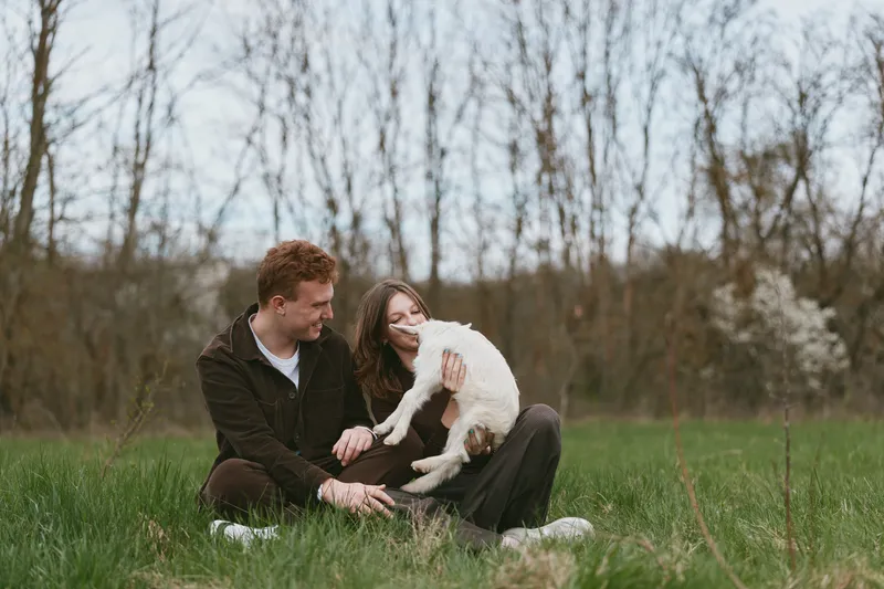 A couple sits on a grassy field enjoying a playful moment with a fluffy dog, conveying casual outdoor lifestyle and companionship in a bright, natural setting, ideal for everyday joy and connection