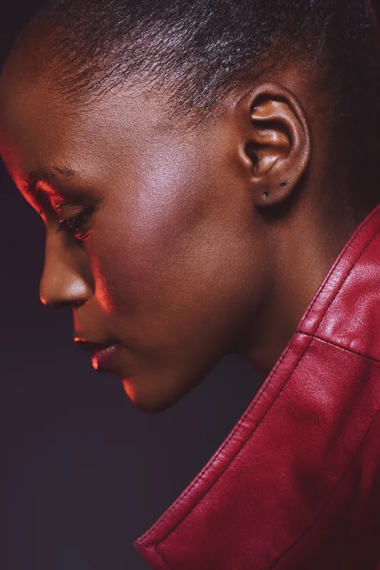 Close-up profile of a young black woman with dark skin wearing a red leather jacket. Studio portrait with dramatic lighting highlights facial features and smooth skin texture.