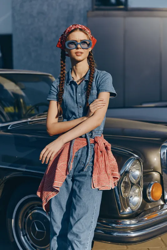 Lifestyle woman in modern boho-western denim street style wearing sunglasses with braided hair and red bandana, standing by vintage car under natural daylight.