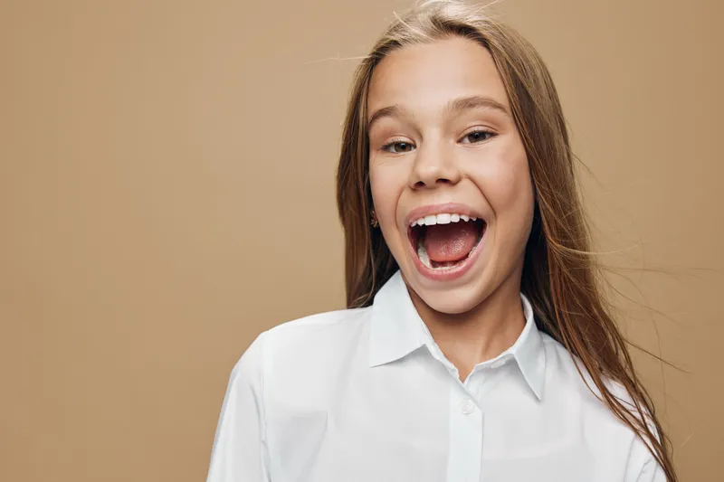 Girl laughing in white shirt with open mouth, long hair and natural expression on beige background, portrait of happy female child with joyful smile.