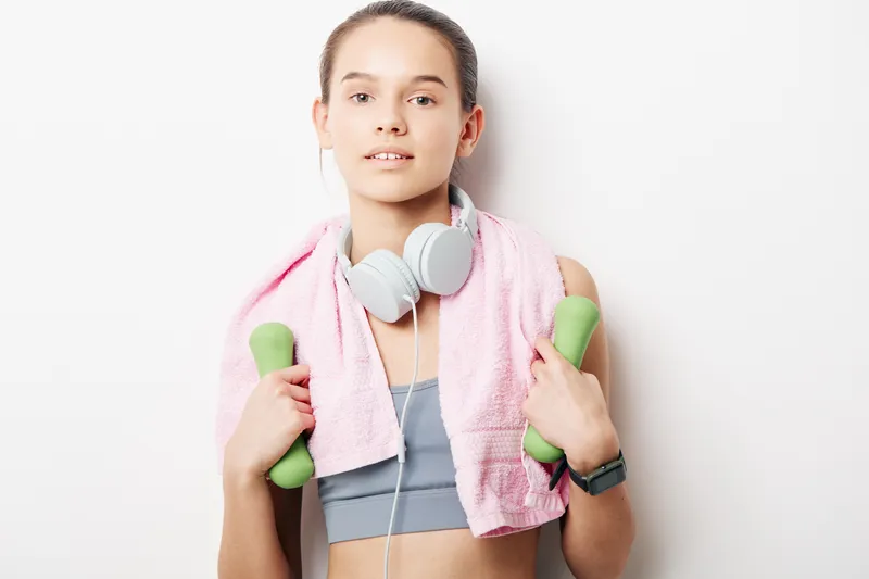 Fitness woman lifting dumbbells after a workout, wearing a grey sports bra and top, with a pink towel around her neck and headphones, standing in a bright gym.