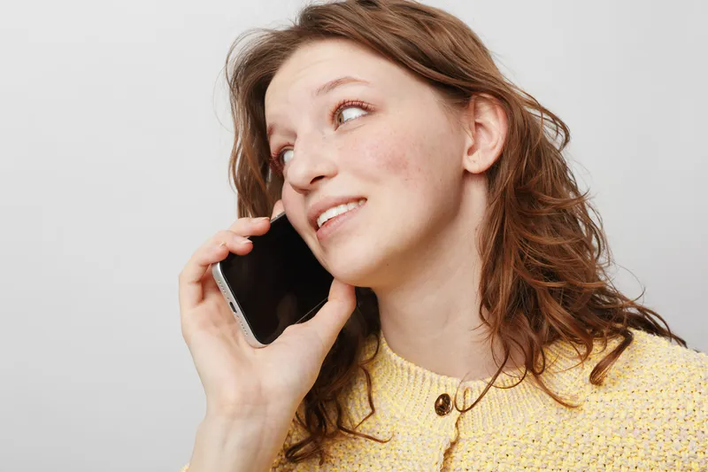 Young woman with curly hair wearing yellow sweater talks on smartphone, smiling and looking away. Bright studio background. Concept of communication, technology, and casual lifestyle.