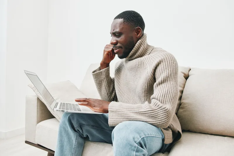 Pensive African man sitting on a couch working on a laptop in a cozy sweater, showcasing deep concentration and thoughtfulness while surrounded by a bright, minimalistic interior