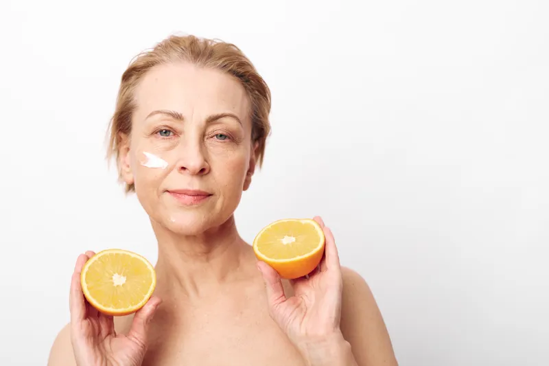 Mature woman with short hair holds two halves of orange, applies natural cream on face. Concept of skincare, healthy lifestyle, and natural beauty in minimalistic studio setting.