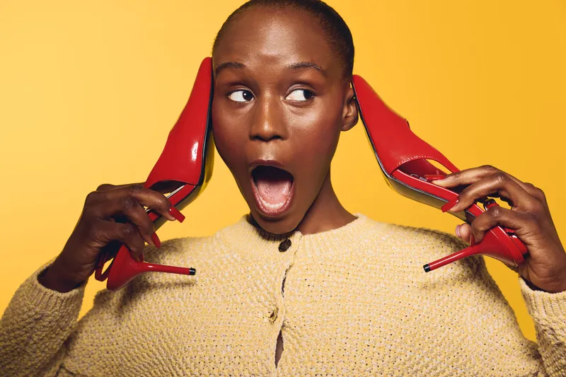 Surprised African woman with dark skin holds red high heels near ears on yellow background. Concept for fashion, beauty, expression, and colorful lifestyle photography.