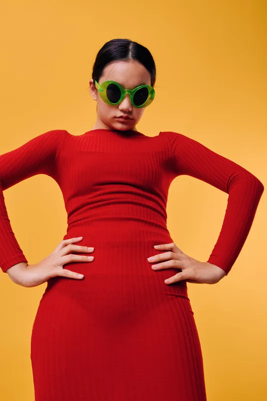 Woman wearing red dress posing with hands on hips and green sunglasses, against bright yellow background. Fashionable model with confident expression and stylish casual look.