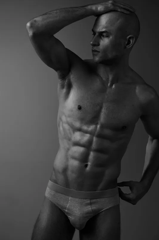 Black and white image of a muscular man striking a confident pose, showcasing his strength and body expression through dramatic lighting and shadow play.