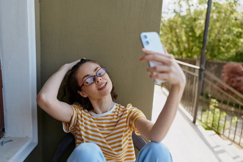 Cheerful woman on a sunny balcony, posing for a selfie, wearing a striped shirt and glasses, relaxed lifestyle moment with natural light and greenery in the background, expressive smile