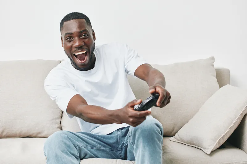 Happy young man playing video games on a couch, wearing a casual outfit, showcasing excitement and joy in a relaxing indoor setting