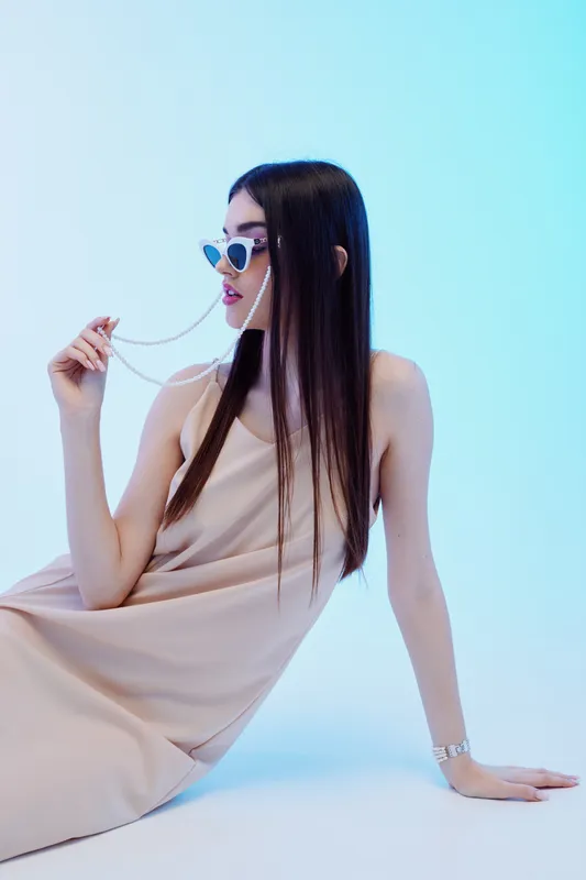 Young woman in beige dress wearing heart-shaped sunglasses and holding a necklace chain. Studio shot with soft blue background. Concept for beauty, fashion, and style.