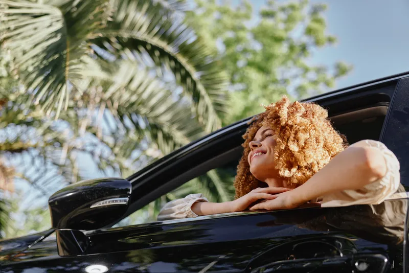 Happy woman with curly hair enjoying a sunny day leaning out of black car window surrounded by green palm leaves and bright blue sky.