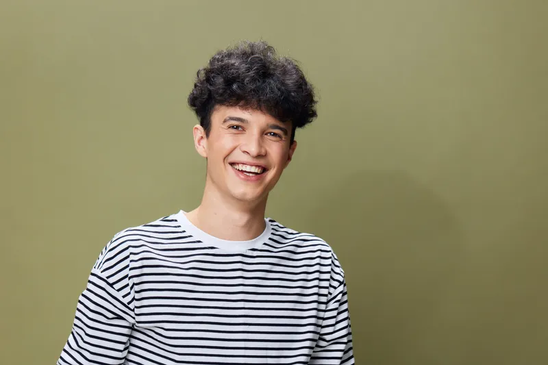 smiling young man in striped shirt with curly hair standing against olive background, expressing happiness and casual style, cheerful and relaxed mood