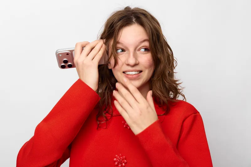 Young woman in red sweater holds smartphone near ear and looks away with surprised expression on white background. Concept of communication, technology, and unexpected news.