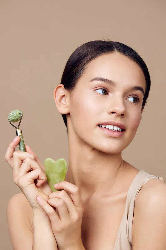 Young woman with jade roller and gua sha stone demonstrating a natural skincare routine, showcasing beauty and wellness in a serene, neutral toned background