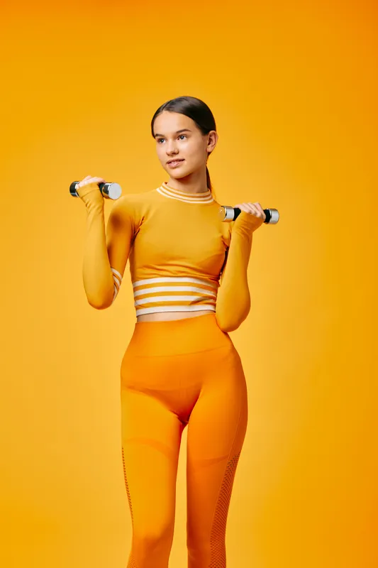 A woman in bright orange fitness wear holds dumbbells in a vivid studio, showcasing strength and balance during a dynamic workout. Bold color and focused expression define a powerful fitness portrait.