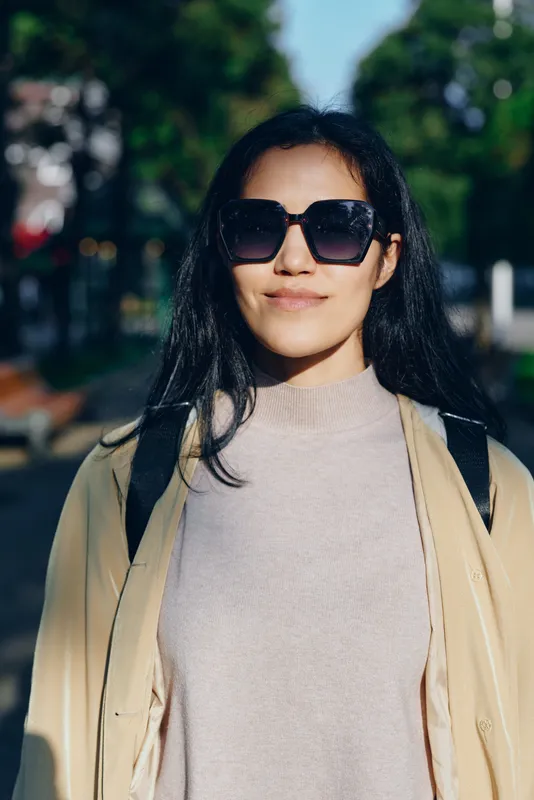 Woman wearing sunglasses, beige coat and knit sweater outdoors on a sunny day, casual fashion vibe, confident pose, urban park setting with trees and soft light.