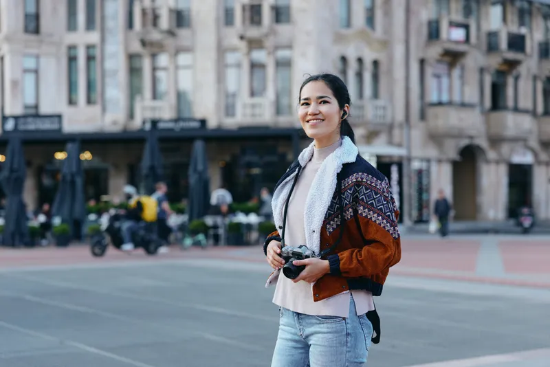 Urban street scene with a smiling woman holding a camera, dressed in casual denim and a knit jacket, ready to capture city moments in an open plaza backdrop and busy storefronts nearby