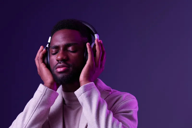 Man enjoying music with headphones, closed eyes, dressed in stylish outfit, against purple gradient background, conveying relaxation and joy