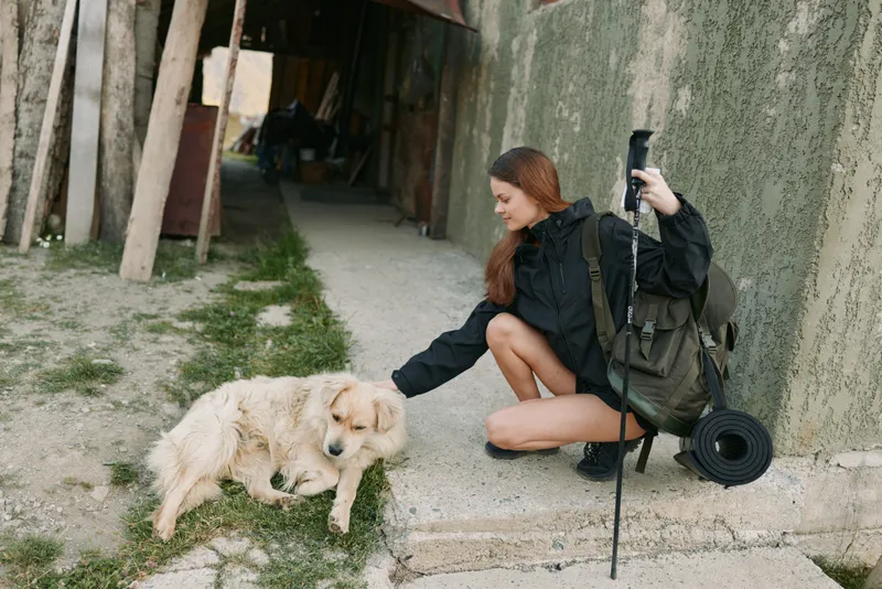 A thoughtful traveler kneels to pat a friendly dog by a rough concrete alley, carrying a hiking backpack and a rolled mat, ready for an outdoor adventure.