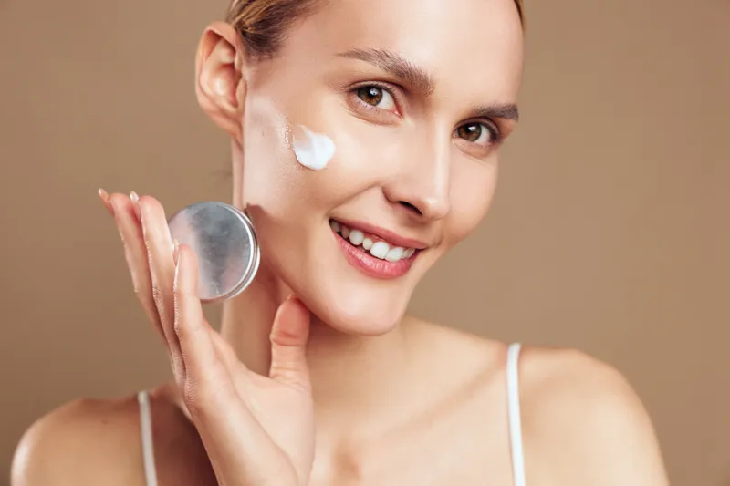 A young woman in a white tank top applies facial cream with a small puff, smiling at the camera in a soft studio setting, illustrating a skincare routine and self care.