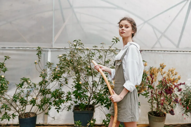Female model in light summer outfit standing in greenhouse, holding gardening tool, surrounded by plants, conveying serene and natural atmosphere