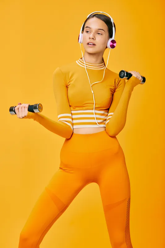 Young woman fitness portrait in a bright orange matching activewear set, holding small dumbbells and wearing headphones against a seamless orange studio background in sporty pose.